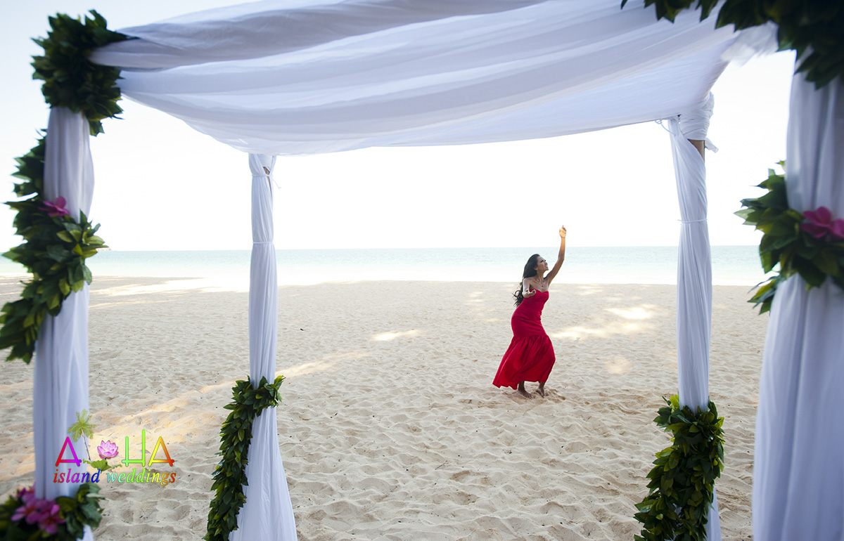 Hula dancer in a red dress dancing on the sand, seen through a white fabric gazebo wrapped in green leaves